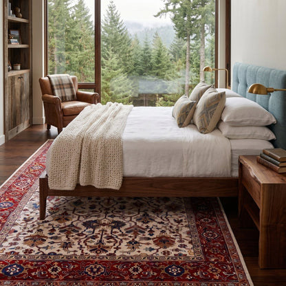 A styled bedroom scene featuring a wooden bed with white linens, patterned pillows, a cream knit throw, and a large red and beige Persian-style area rug. The room has tall windows overlooking a forest, a leather armchair, a nightstand, and a blue upholstered headboard.