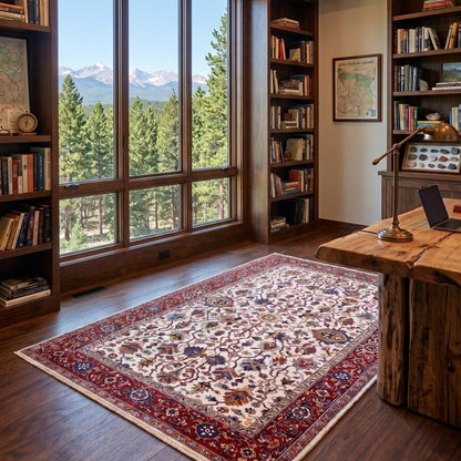 A traditional oriental area rug with a red border and cream floral pattern is displayed on a dark wood floor in a cozy, book-filled home office with a large window overlooking pine trees and mountains.