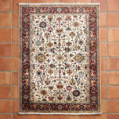 An ornate rectangular area rug with a cream center field and a dark red border, displayed flat against a rustic terracotta brick wall.