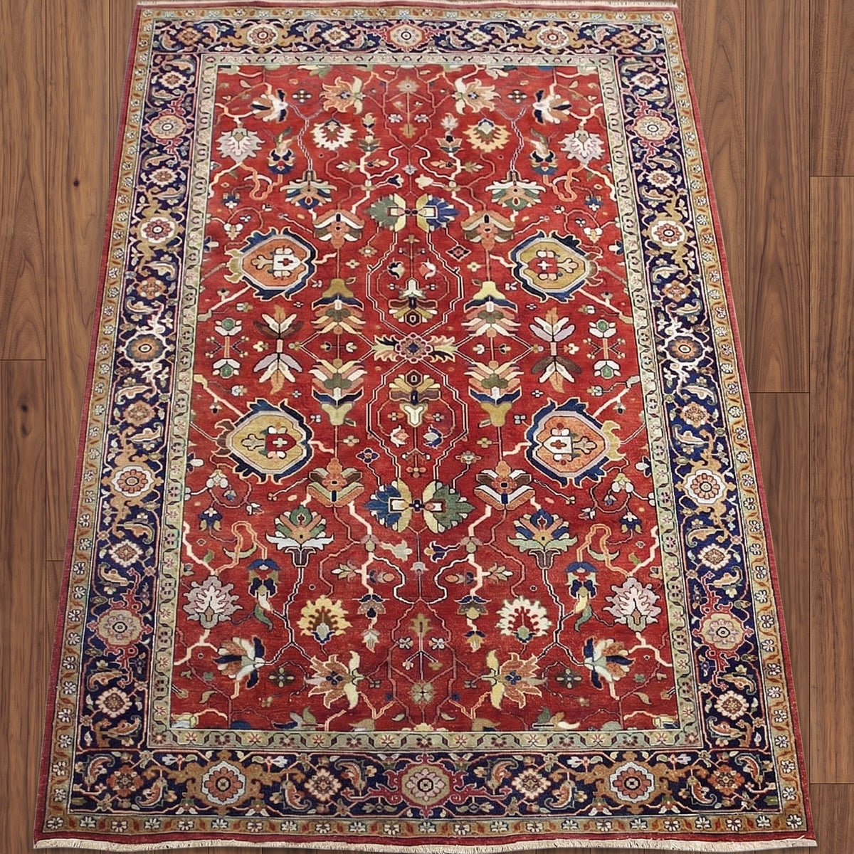 A traditional Persian-style area rug with a red field, navy blue borders, and intricate floral motifs in beige, green, and blue, laid flat on a brown wooden floor.