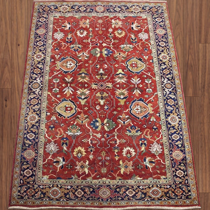 A traditional Persian-style area rug with a red field, navy blue borders, and intricate floral motifs in beige, green, and blue, laid flat on a brown wooden floor.